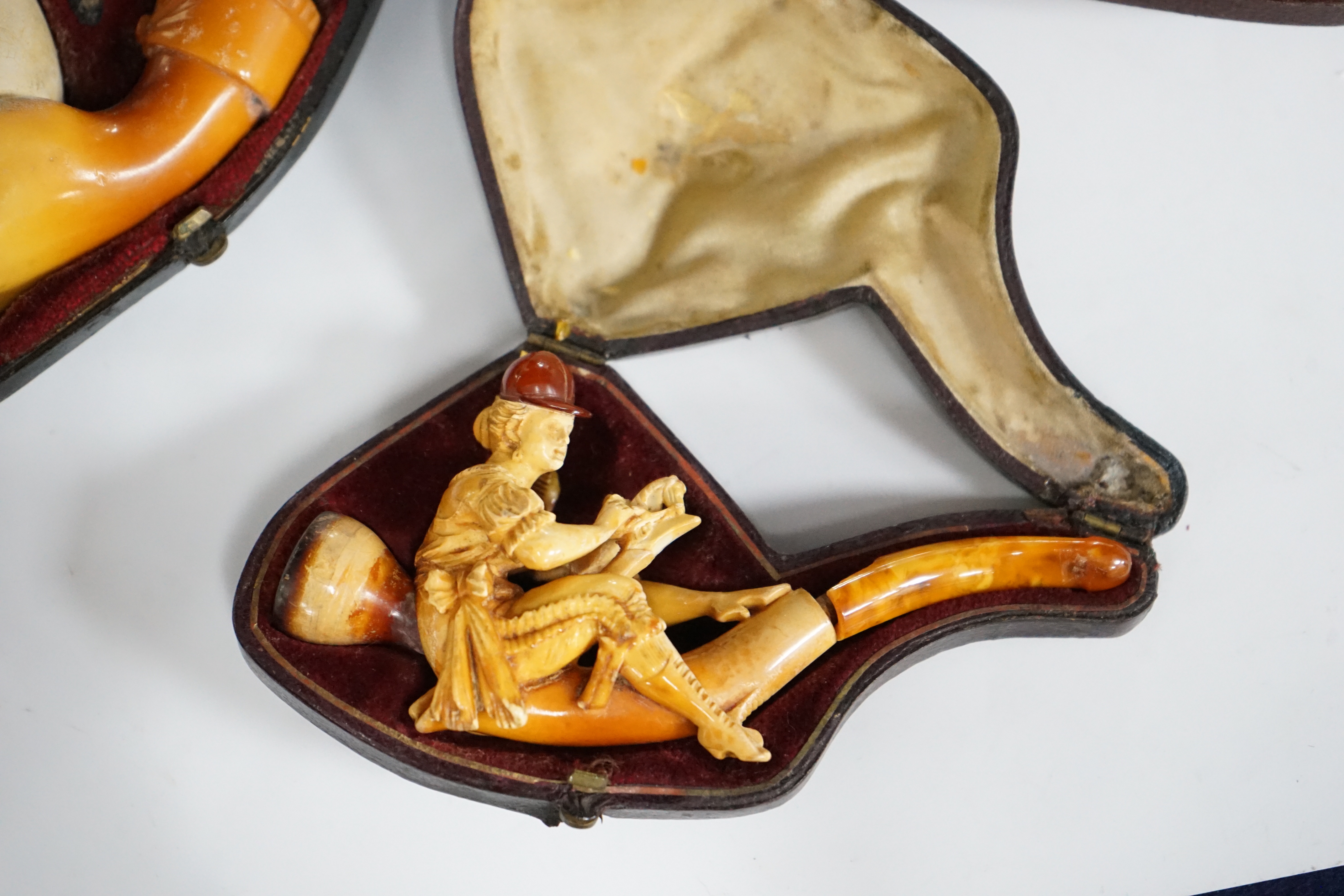 Three cased novelty Meerschaum pipes and pipe bowls, all with amber mouth pieces, one of a horses and cowboy, another a model of a hand and the other of a scantily clad lady wearing a riding cap, largest 17 cm long